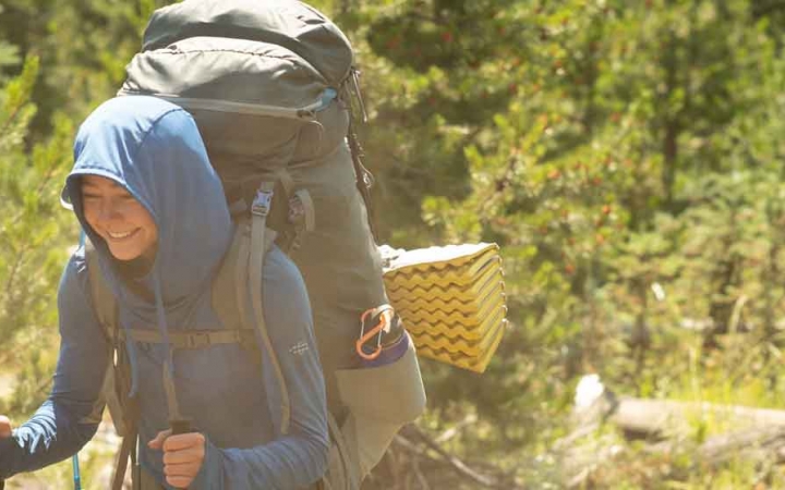 a student carrying a backpack smiles as they hike among trees on an outward bound course 